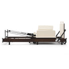 Foldable Pilates Reformer Flexi, dark wood, milky white upholstery – side view with sitting box